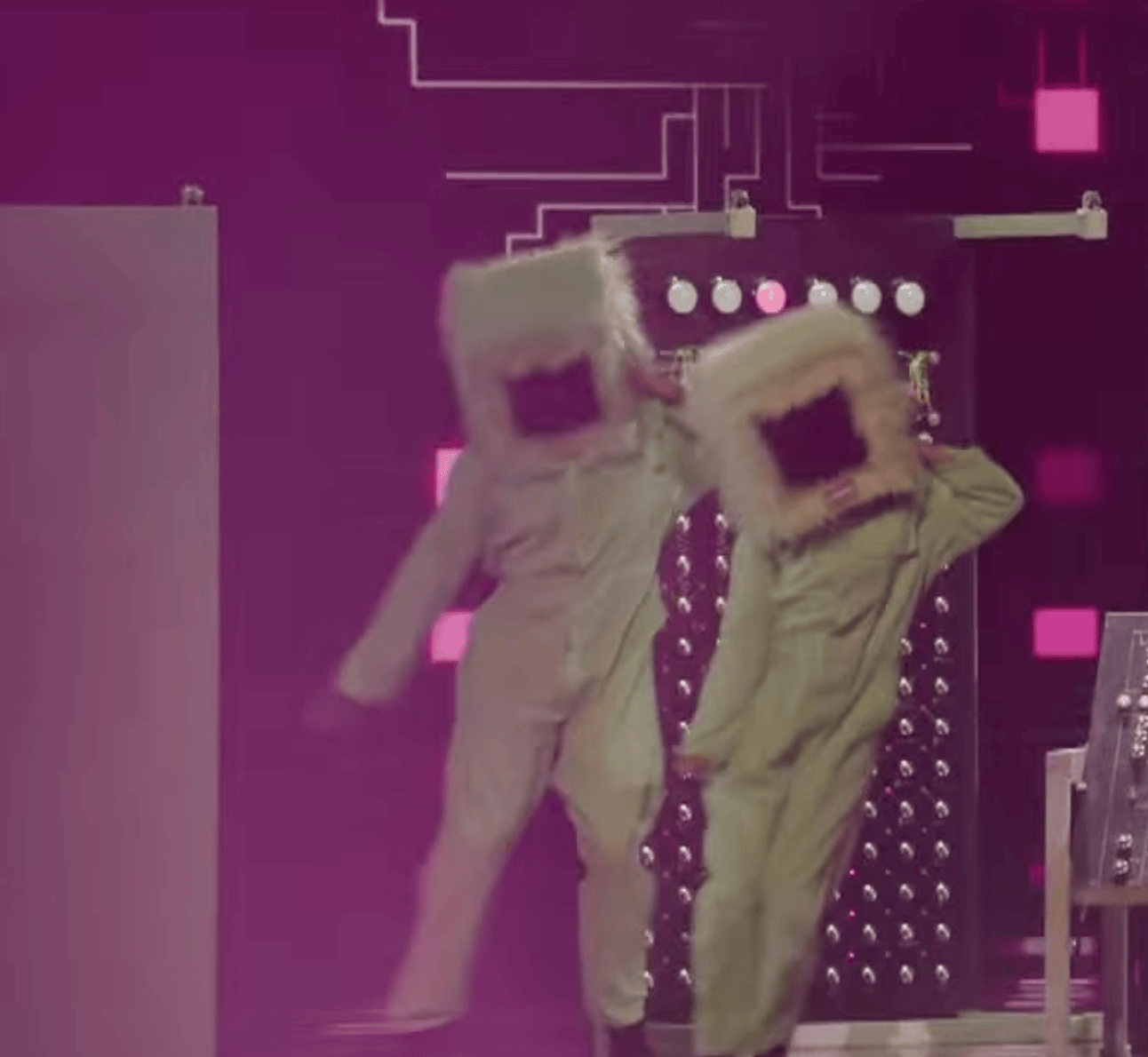Two people wearing large, rectangular masks and light-colored outfits are dancing energetically in front of a futuristic backdrop.